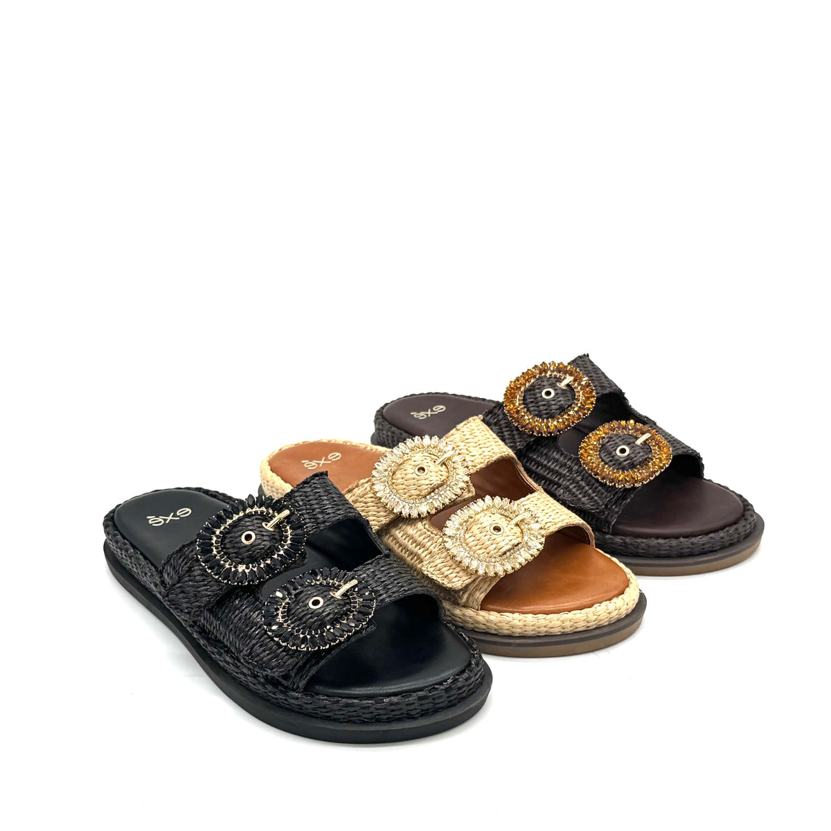 Three women's slide sandals arranged on a white background, each with a large decorative oval buckle embellished with rhinestones. The sandals come in black, tan/beige, and dark brown espadrille-style uppers with braided trim.