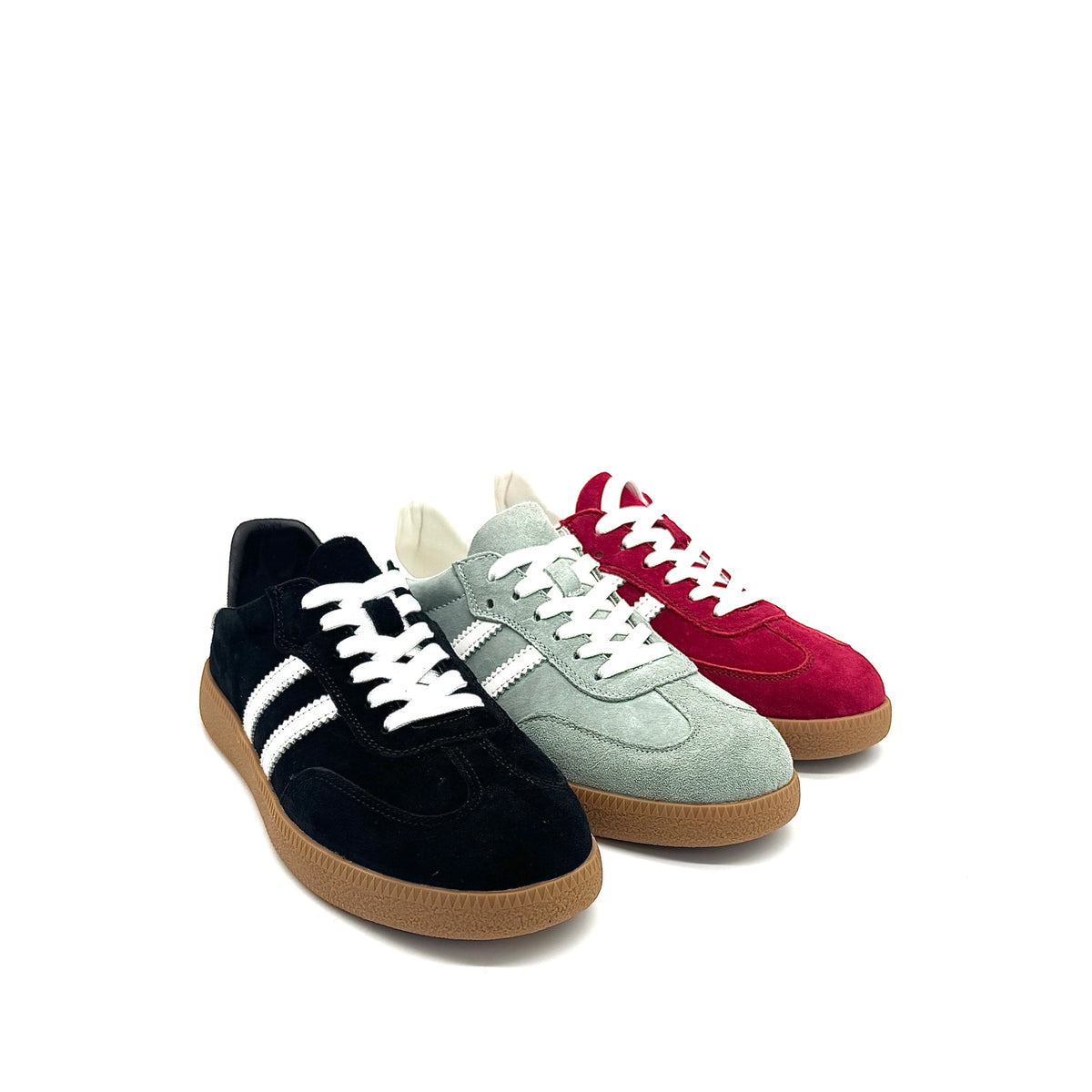 Three low-top suede sneakers arranged in a fan shape on a white background: a light sage green pair, a black pair, and a deep red pair, each with white laces and white side stripes, plus gum soles.