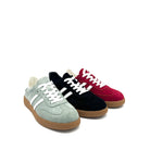 Three low-top suede sneakers arranged in a fan shape on a white background: a light sage green pair, a black pair, and a deep red pair, each with white laces and white side stripes, plus gum soles.