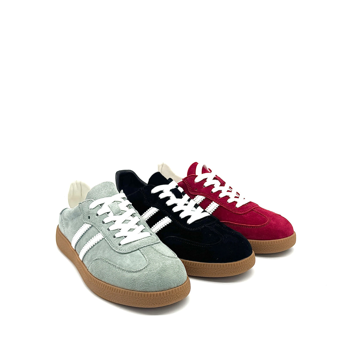 Three low-top suede sneakers arranged in a fan shape on a white background: a light sage green pair, a black pair, and a deep red pair, each with white laces and white side stripes, plus gum soles.