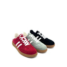 Three low-top suede sneakers arranged in a fan shape on a white background: a light sage green pair, a black pair, and a deep red pair, each with white laces and white side stripes, plus gum soles.