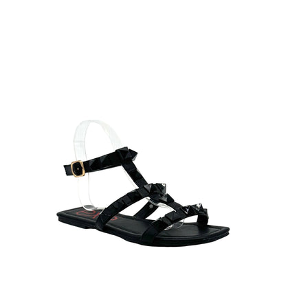 Black: EXE 2269-2781 pyramid stud T-strap flat sandal in black with gold-tone ankle buckle and square toe, side view