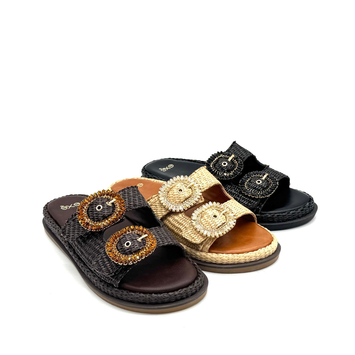 Three women's slide sandals with braided jute trim and large decorative oval buckle details, shown on a white background. The sandals are in black, tan, and dark brown.